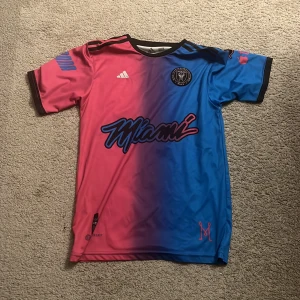 Inter Miami kortärmad fotbollströja - Snygg Inter Miami fotbollströja från Adidas med cool färgövergång mellan rosa och blått. Tröjan har svart krage och muddar, klubbmärke på bröstet och Miami-tryck framtill. Tillverkad i lätt polyester som andas, perfekt för match eller träning.