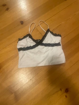 Vit linnetopp med svart spets från Sister - Säljer en vit linnetopp från Sister i storlek M. Toppen har tunna axelband och är dekorerad med svart spets längs halsringningen och under bysten. Snygg croppad passform som ger en trendig vibe. Perfekt för dig som gillar kontraster och detaljer.
