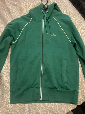 Grön hoodie från GANT med dragkedja - Snygg grön hoodie från GANT med vit piping längs axlarna och hel dragkedja framtill. Tröjan har huva med snörning och två fickor på sidorna. Perfekt för chill dagar och har en stilren look med diskret logga på bröstet.