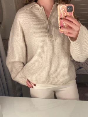 Beige ribbad stickad tröja med krage - Säljer en beige stickad tröja med ribbad struktur och krage. Tröjan har tre knappar framtill och en loose passform som ger en chill vibe. Perfekt för lager-på-lager och passar till allt. Långärmad och mjuk, riktigt skön att ha på sig.
