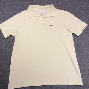 Ljusgul Lacoste pikétröja i bomull - Snygg ljusgul pikétröja från Lacoste med klassisk krage och korta ärmar. Tröjan har två knappar vid halsen och den ikoniska krokodilloggan broderad på bröstet. Tillverkad i mjuk bomull som är skön mot huden och passar perfekt till sommaren.