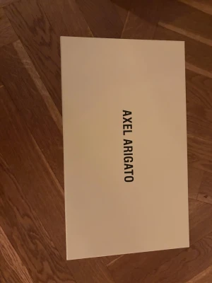 Svarta sneakers från Axel Arigato - Svarta slip-on sneakers från Axel Arigato med vit platt sula och rund tå. Skorna har en stilren design med bred elastisk rem över foten och logga på plösen samt undersidan. Ovandelen är i mocka och sulan i gummi, perfekt för dig som gillar minimalistisk streetstil.