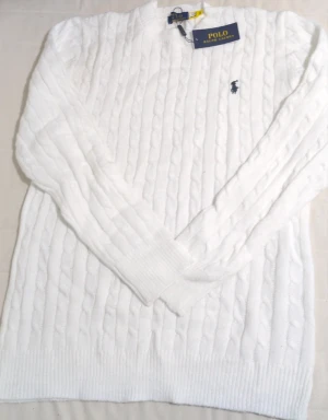 Vit kabelstickad tröja Polo Ralph Lauren - Vit kabelstickad tröja från Polo Ralph Lauren med klassisk rund hals och broderad logga på bröstet. Tröjan har långa ärmar och ribbade muddar vid ärmslut och nederkant. Perfekt för dig som gillar clean och tidlös stil.