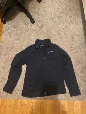 Svart fleecejacka från Patagonia - Mysig svart fleecejacka från Patagonia med hel dragkedja framtill och hög krage. Jackan har två sidofickor med dragkedja och en bröstficka med logotyp. Perfekt för kyliga dagar och enkel att slänga på sig när du är på språng.