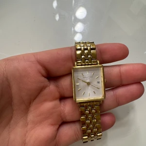 Guldfärgad fyrkantig klocka Rosefield - Snygg guldfärgad klocka från Rosefield med fyrkantigt ur och länkarmband i metall. Urtavlan är vit med minimalistiska guldmarkeringar och datumvisning. Perfekt accessoar för dig som gillar stilrena detaljer och klassisk design.