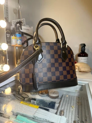 Louis Vuitton Damier handväska brun - Snygg handväska från Louis Vuitton i klassiskt Damier-mönster. Väskan är brun med mörkbruna detaljer, guldfärgade metalldetaljer och dragkedja. Tillverkad i skinn och har både handtag och axelrem. Perfekt för dig som vill ha en ikonisk och stilren accessoar. Vid snabb affär kan ja sänka priset lite grann 