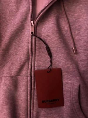 Grå hoodie från Burberry med dragkedja - Snygg grå hoodie från Burberry med klassisk huva och snörning. Insidan av huvan har det ikoniska Burberry-rutiga mönstret i beige och rött. Tröjan är tillverkad i 100% bomull och har praktisk dragkedja framtill. Perfekt för dig som gillar stilrena och exklusiva plagg. Helt ny med prislapp