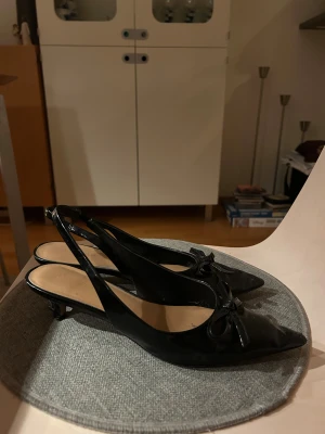 Svarta klackar med rosett och spetsig tå - Säljer ett par svarta klackar med elegant spetsig tå och söt rosettdetalj framtill. Skorna har låg klack och öppen häl, vilket ger en stilren och trendig look. Tillverkade i blankt skinnmaterial som ger en lyxig känsla.