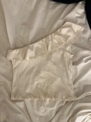 Vit oneshoulder topp från H&M Divided - Säljer en vit oneshoulder topp från H&M Divided i storlek S. Toppen har en snygg volang längs halsringningen och är tillverkad i ett lätt, strukturerat tyg. Perfekt för dig som gillar en feminin och trendig stil.