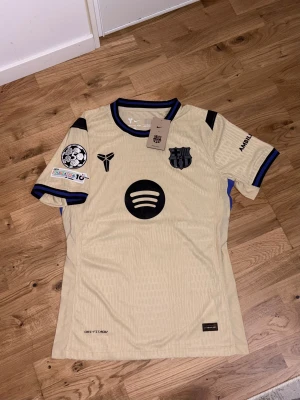 FC Barcelona Away Jersey 25/26 – Rafinha #11 – Size M/ L - Säljer en beige FC Barcelona fotbollströja från Nike med Raphinha och nummer 11 på ryggen. Tröjan har svarta och blå detaljer, korta ärmar och flera tryck, bland annat Spotify-loggan och UNHCR. Materialet är lätt och andas, perfekt för match eller träning.