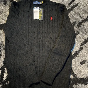 Svart stickad tröja från Polo Ralph Lauren - Svart kabelstickad tröja från Polo Ralph Lauren med klassisk rund hals och rött broderat logomärke på bröstet. Tröjan har långa ärmar och ribbade muddar vid ärmslut och nederkant. Perfekt för dig som gillar stilren och tidlös design.