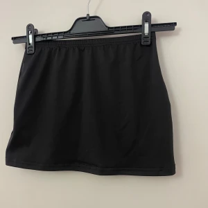 Svart kortkjol med enkel design - Snygg svart kortkjol med rak passform och elastisk midja. Kjolen är stilren och passar perfekt till en trendig streetstyle-look, vardagligt bruk samt klä upp mer festligt! Materialet känns lätt och bekvämt, vilket gör den enkel att matcha med olika toppar.