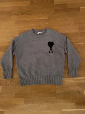 Grå stickad tröja från Ami - Snygg grå stickad tröja från Ami med svart hjärta och A broderat på bröstet. Tröjan har rund hals, ribbade muddar och nederkant samt långärmad passform. Perfekt för dig som gillar stilrena detaljer och vill ha något som sticker ut lite.