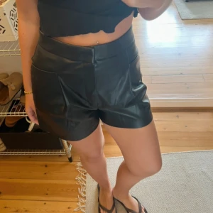 Svarta högmidjade skinnshorts - Snygga svarta shorts i skinnimitation med hög midja och bred linning. De har två framfickor och diskreta bakfickor, samt stängs med dragkedja framtill. Passformen är loose och modellen är lite A-linjeformad, vilket ger en cool och trendig vibe.