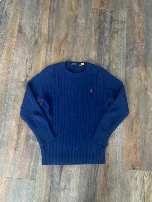 Marinblå stickad tröja Polo Ralph Lauren - Snygg marinblå stickad tröja från Polo Ralph Lauren i storlek S. Tröjan har ett klassiskt kabelstickat mönster och en röd broderad logga på bröstet. Rund hals och långa ärmar med ribbade muddar. Perfekt för dig som gillar stilrena och tidlösa plagg.