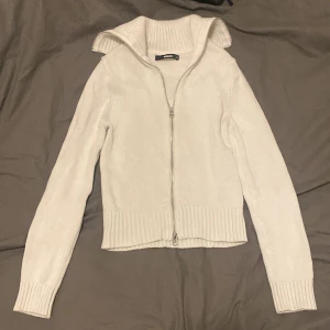 Vit stickad kofta från Bik Bok - Säljer en vit stickad kofta från Bik Bok i storlek XS. Koftan har en bred, ribbad krage och hel dragkedja framtill. Modellen är långärmad med ribbade muddar och nederkant, perfekt för lager-på-lager under kyliga dagar.