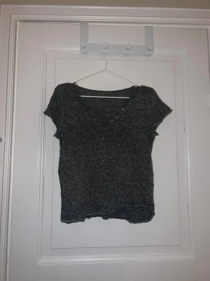 Mörkgrå ribbad topp med v-ringning - Säljer en mörkgrå ribbad topp med korta ärmar och snygg v-ringning framtill. Toppen har fyra små knappar längs halsringningen som ger en cool detalj. Materialet är mjukt och stretchigt.