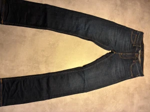 Mörkblå jeans från Nudie  27/32 - Snygga mörkblå jeans från Nudie  i klassisk femficksmodell med orangea sömmar och läderpatch bak. Modellen har smal passform och raka ben, tillverkade i slitstarkt jeanstyg. Perfekt för dig som gillar stilrena och tidlösa jeans. Är i ny skick
