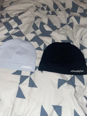 2 Satynmössor i svart och silver 300kr per mössa - Två stilrena mössor från Satyn, en i svart och en i ljus silver. Utsidan är slät och enkel med broderad logga, insidan är fodrad med glansig satin för extra komfort och minskat friss. Perfekt för dig som vill skydda håret och ändå se clean ut.