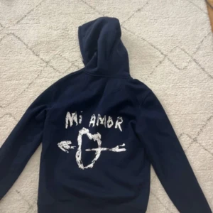 Mörkblå hoodie med Mi Amor-tryck - Snygg mörkblå hoodie med coolt vitt tryck på ryggen där det står 'Mi Amor' och en pil genom ett hjärta. Hoodien har huva och långa ärmar, perfekt för dig som gillar streetstyle och vill sticka ut med ett unikt motiv. Kostar 1500 ny!