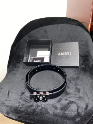 Svart skärp från Amiri med metallspänne - Säljer ett svart bälte från Amiri med grov textur i skinn och ett unikt metallspänne med Amiri-logga i silver och svart. Bältet kommer med originalask och certifikat. Perfekt för dig som vill ha en stilren och lyxig detalj till outfiten.