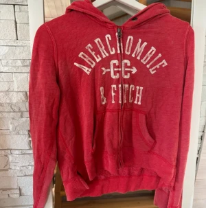 Röd hoodie Abercrombie & Fitch M - Snygg röd hoodie från Abercrombie & Fitch i storlek M. Tröjan har vit logotyp framtill, huva med snörning och en praktisk känguruficka. Dragkedja hela vägen och mjukt material som känns skönt mot huden. Perfekt för chill dagar och streetstyle.