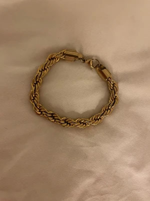 Cordell armband  - Snyggt flätat armband i guldfärgad metall med robust karbinlås. Armbandet är cirka 19 cm långt och har en cool, tvinnad design som ger en exklusiv känsla. Perfekt för dig som gillar statement-smycken och vill ha något som sticker ut.
