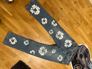 Levi's 501 Denim Tears jeans med blommor - Säljer ett par Levi's 501 Denim Tears jeans i klassisk blå tvätt med vita blommiga tryck över hela benen. Jeansen har rak passform, fem fickor och knappgylf. Märket syns tydligt på bakfickan och patchen. Perfekt för dig som vill sticka ut med unik stil.