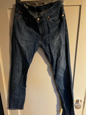 Levi's 511 blå jeans W33 L34 - Säljer ett par klassiska Levi's 511 jeans i blå denim. Modellen är slim fit med raka ben och fem fickor. Jeansen har snygga kontrastsömmar, läderpatch bak och den ikoniska röda Levi's-taggen. Perfekta för dig som gillar en tidlös och stilren look.