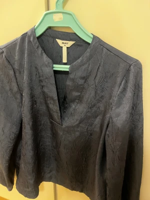 Mörkgrå blus från Object - Säljer en mörkgrå blus från Object med snyggt skrynklat tyg och v-ringad hals. Blusen har långa ärmar och en rak, något lös passform. Perfekt för dig som gillar stilrena plagg med en twist.