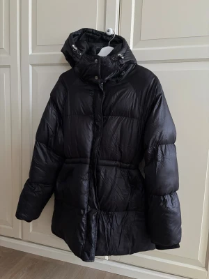 Svart dunjacka  - En svart medellång dunjacka från Ellos, storlek 38/40. Möjlighet att dra åt i midjan och muddar i armarna. 