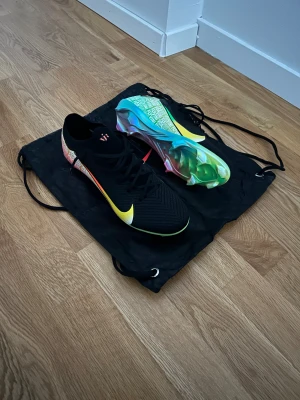 Vini Jr Nike Fotbollskor - Säljer ett par Nike fotbollsskor i storlek 43. Skorna är svarta med en stor gul swoosh och har färgglada detaljer på hälen och sulan. Ovandelen är i syntetmaterial med snörning och platt sula med dobbar. Sulan har ett coolt mönster och text. Perfekt för dig som vill sticka ut på planen.
