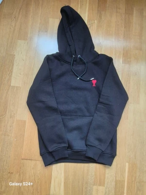 Svart hoodie från Ami Paris med rött hjärta - Svart hoodie från Ami Paris med klassisk huva och snörning. På bröstet finns ett broderat rött hjärta med bokstaven A. Tröjan har en stor magficka och ribbade muddar vid ärmslut och nederkant. Perfekt för chill dagar.