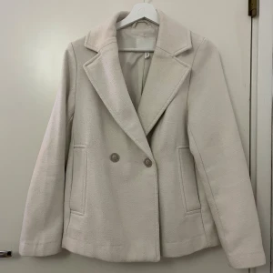 Beige kort kappa från H&M XS - Snygg beige jacka/kappa från H&M i storlek XS. Jackan har klassisk krage, dubbelknäppning med två knappar och två fickor fram. Materialet är mjukt och har en ull-liknande känsla, perfekt för kyliga dagar. Stilren och enkel design som passar till många outfits.