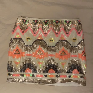 Mönstrad paljettkjol - Snygg kortkjol med färgglatt mönster och glittriga paljetter. Kjolen har en mix av rosa, guld och orange nyanser som ger en festlig look. Perfekt för att sticka ut med stil!