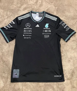 Mercedes AMG Petronas fotbollströja - Snygg svart Mercedes AMG Petronas Formula One Team fotbollströja från Adidas. Tröjan har korta ärmar, vit och turkos detalj på krage och ärmslut samt flera sponsorer tryckta på bröstet och ryggen. Tillverkad i lätt och ventilerande polyester med Aeroready-teknologi.