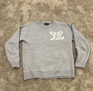 Grå stickad Louis Vuitton tröja - Snygg grå stickad tröja från Louis Vuitton med stort vitt LV-logo på bröstet. Tröjan har rund hals, ribbade muddar och är långärmad. Perfekt för dig som gillar exklusiva märken och vill ha en clean look med en touch av lyx.