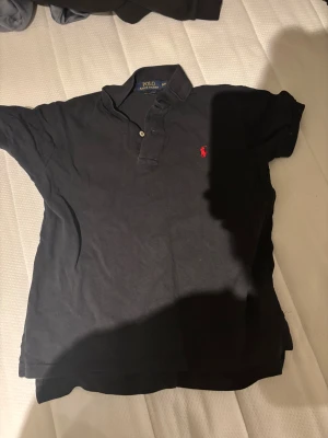 Svart pikétröja från Polo Ralph Lauren - Klassisk svart pikétröja från Polo Ralph Lauren med röd broderad logga på bröstet. Tröjan har korta ärmar, krage och två knappar vid halsen. Perfekt för en clean och stilren look.