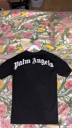 Svart Palm Angels t-shirt med tryck - Svart t-shirt från Palm Angels med stort vitt logotryck på ryggen och mindre tryck vid halsen fram. Klassisk rund hals och korta ärmar. T-shirten har en clean och stilren look, perfekt för dig som gillar streetwear och vill sticka ut med ett ikoniskt märke. 