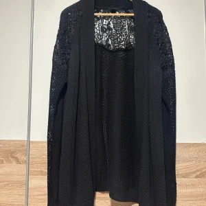 Svart stickad kofta med spetsdetaljer - Snygg svart stickad kofta med öppet framtill och detaljerade spetsinslag på axlar och övre rygg. Perfekt för dig som gillar en chill och stilren look med lite extra edge. Långärmad och mjuk, passar till många outfits.