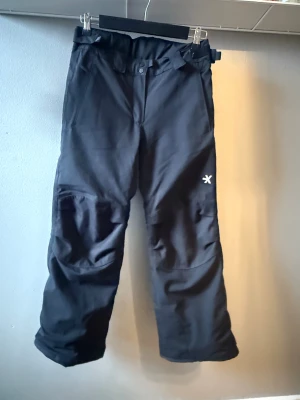 Svarta täckbyxor med förstärkta knän - Svarta skidbyxor från Everest med förstärkta partier vid knän och benslut, justerbar midja med kardborre och knappstängning. Byxorna har snölås i bensluten och är gjorda i slitstarkt syntetmaterial, perfekta för kalla dagar och vinteraktiviteter.