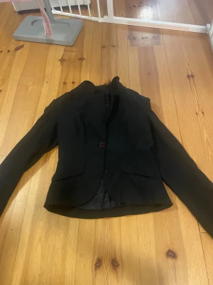 Svart kavaj - Stilren svart kavaj från Vero Moda i storlek 38. Kavajen har en enkel knäppning med en knapp framtill, två fickor med lock och klassisk krage. Perfekt för en snygg och clean look. Materialet är mjukt och har en lätt glansig insida.