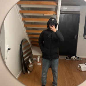 Svart pufferjacka från Sail Racing - Säljer en svart pufferjacka från Sail Racing med huva och broderad logga på både bröstet och huvan. Jackan har dragkedja framtill och flera praktiska fickor. Perfekt för kalla dagar när du vill hålla stilen och samtidigt vara varm. Lite hål runt baksidan av armen men det går att sy ihop matchar avän priset