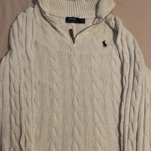 Vit stickad tröja Polo Ralph Lauren - Kabelstickad vit tröja från Polo Ralph Lauren med halv dragkedja och ribbad krage. Klassisk broderad logga på bröstet och snyggt läderdetalj på dragkedjan. Perfekt för lager-på-lager under kyliga dagar. 