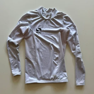 Vit långärmad träningströja Sondico - Vit långärmad träningströja från Sondico med hög krage och tight passform. Perfekt för sport eller gym, tillverkad i stretchigt syntetmaterial som andas. Diskret logga på bröstet och sömmar som ger en sportig look. även kompression shirt.