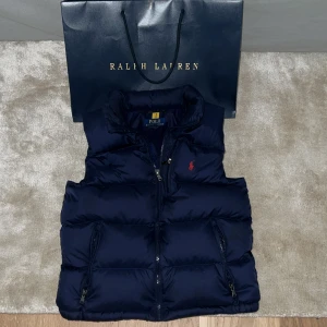 Mörkblå dunväst från Polo Ralph Lauren - Snygg mörkblå dunväst från Polo Ralph Lauren med röd logga på bröstet. Västen är i nyskick och endast använd ett fåtal gånger. Perfekt för dig som gillar grisch stil och vill ha en riktigt fett höstvest. Pris kan diskuteras!