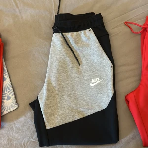 Nike tech byxor - Snygga mjukisbyxor från Nike i svart och ljusgrått med vit logga på benet. Byxorna har dragsko i midjan och är tillverkade i mjukt bomullsmaterial. Perfekta för chill eller träning, med en modern och sportig look.