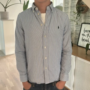 Randig skjorta från Ralph Lauren - Säljer en klassisk blåvit randig skjorta från Ralph Lauren med button-down krage och broderad logga på bröstet. Skjortan har långa ärmar, knappar framtill och är gjord i mjuk bomull. Perfekt för en clean och stilren look.