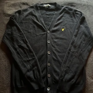 Svart kofta från Lyle & Scott, XS-S - Svart kofta från Lyle & Scott med klassisk v-ringning och knäppning framtill. Broderad gul logga på bröstet. Långärmad och i mjukt material som känns skönt mot huden. Perfekt att slänga över en t-shirt för en clean look.