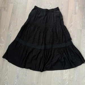 Svart långkjol med spets från KappAhl - Svart långkjol från KappAhl med bred resår i midjan. Kjolen har flera volanger och dekorativa spetsdetaljer som ger en bohemisk vibe. Tillverkad i mjuk bomull som känns skön mot huden. 